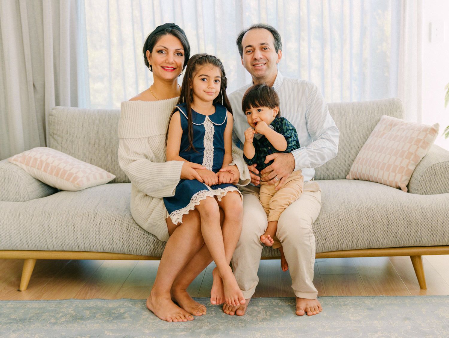 A happy family of four poses together on a light gray sofa in a bright, modern living room.