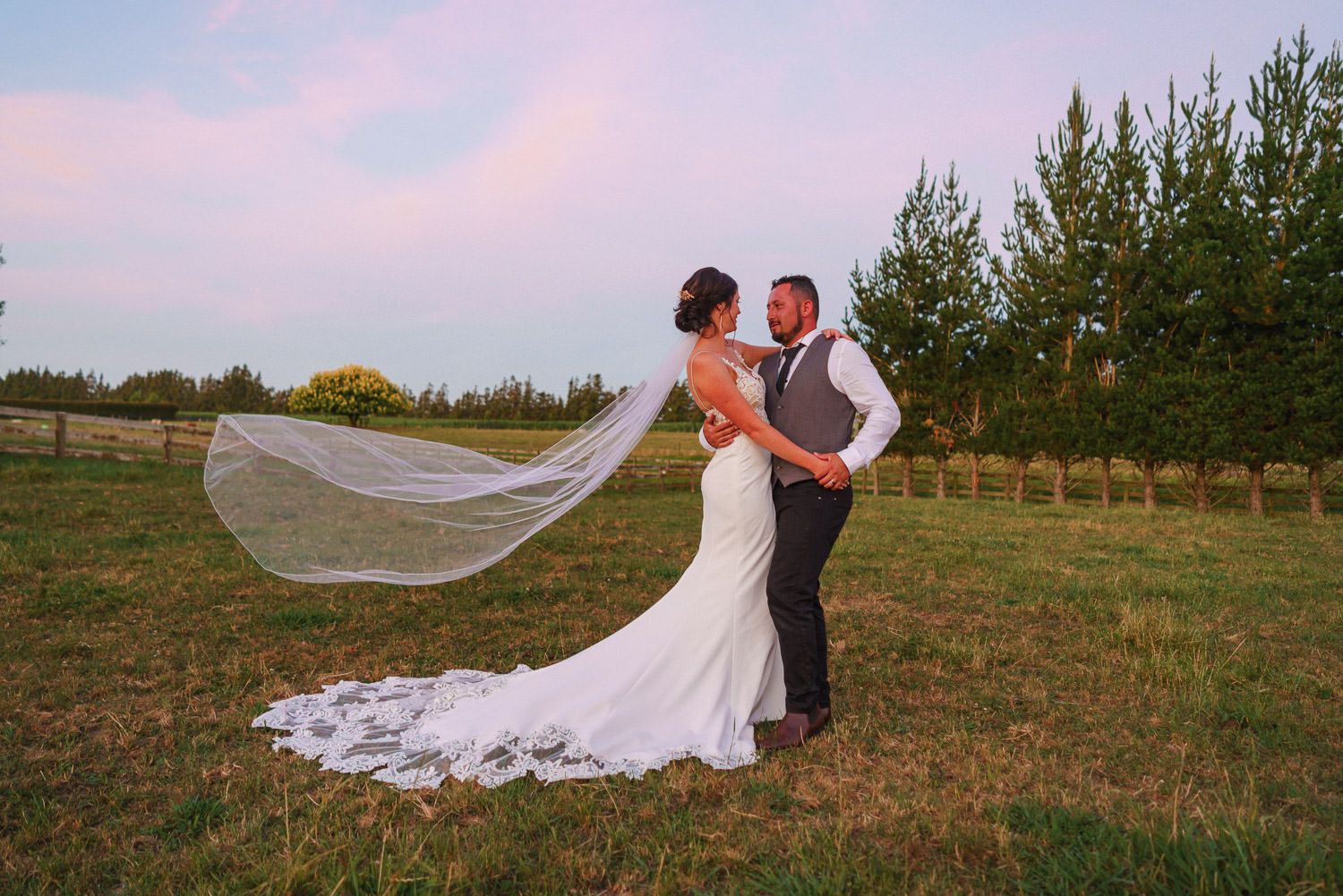 Bride and groom share romantic moment in field, her veil flowing in wind, surrounded by trees at golden hour.