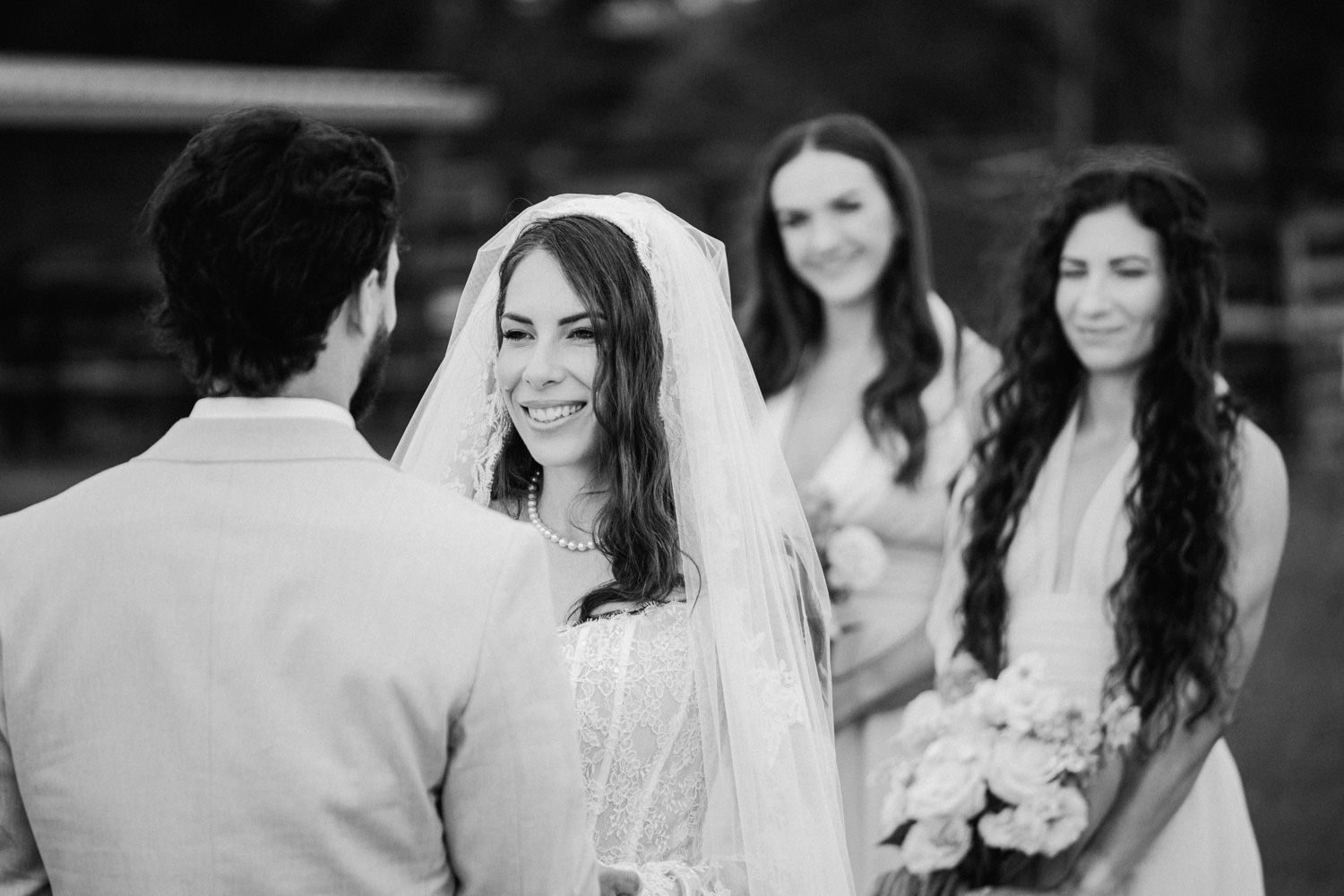 Black and white wedding ceremony photo of smiling bride in veil facing groom with bridesmaids holding bouquets in background.