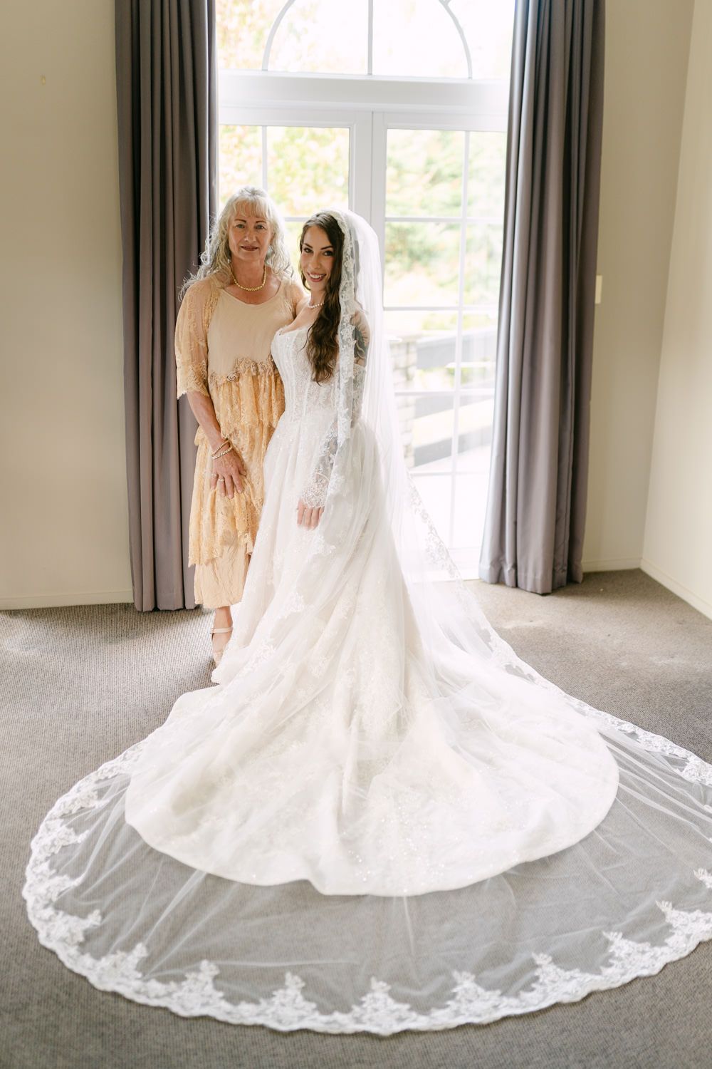 Bride in lace wedding gown with cathedral veil stands with older woman in gold dress by bright window.