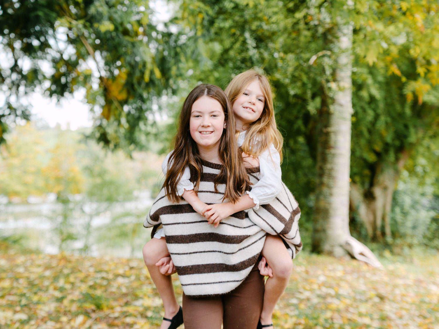 Two young girls smile outdoors in autumn, one giving the other a piggyback ride among colorful fallen leaves.