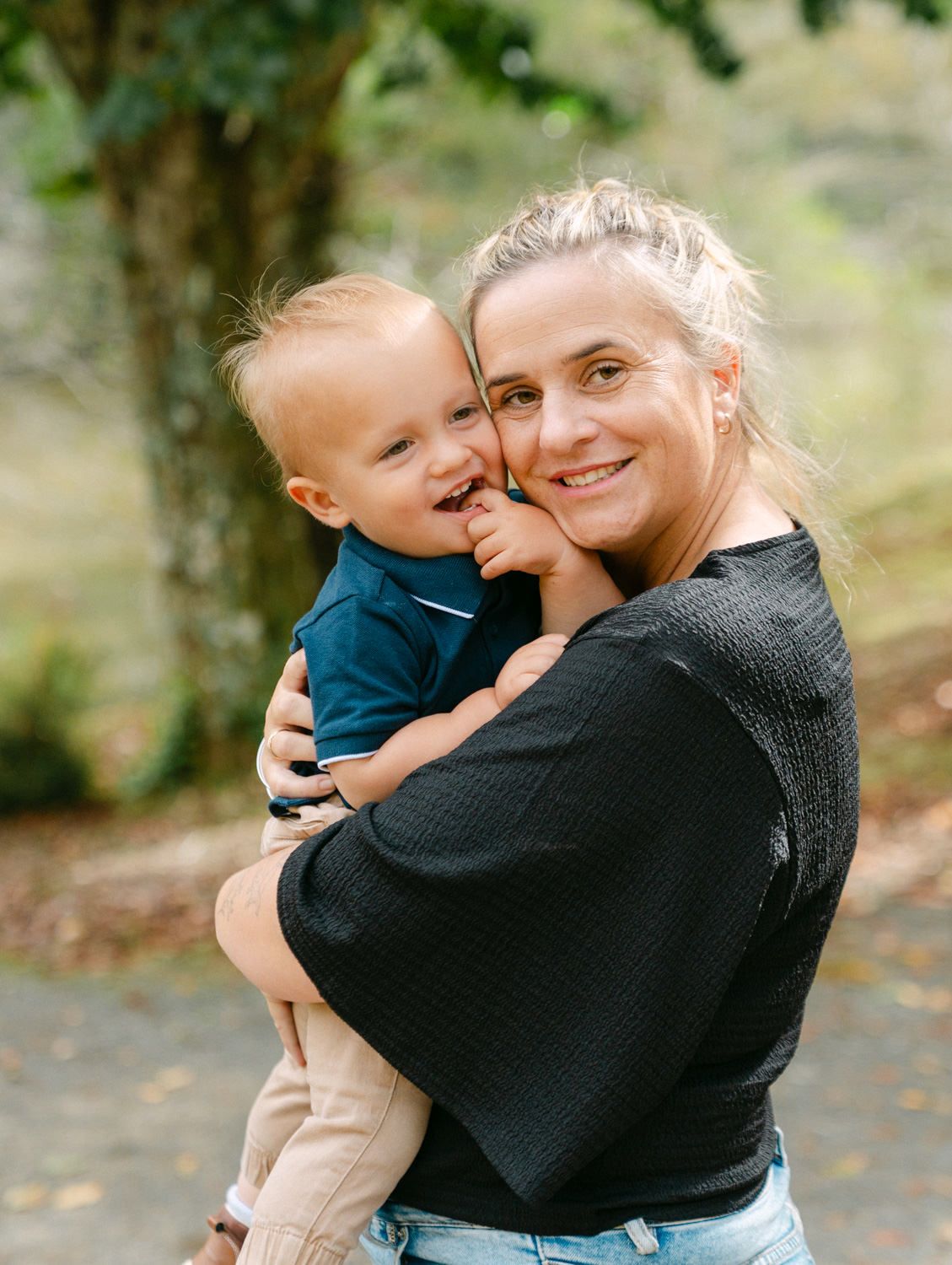 A smiling woman holds a happy toddler boy outdoors in a park setting with autumn leaves on the ground.