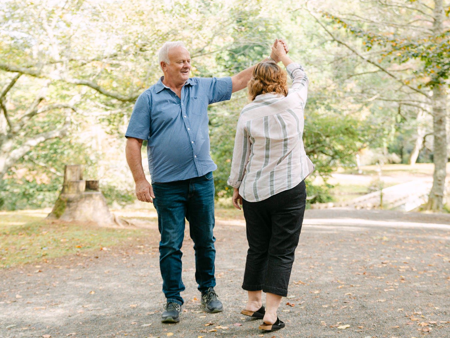 Elderly couple dancing joyfully on a park path surrounded by lush green trees on a sunny day.