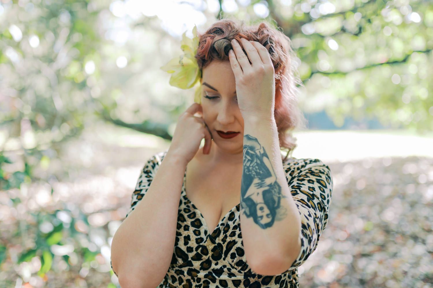 Woman in leopard print dress with hand tattoo poses outdoors among trees, hands raised to her face.