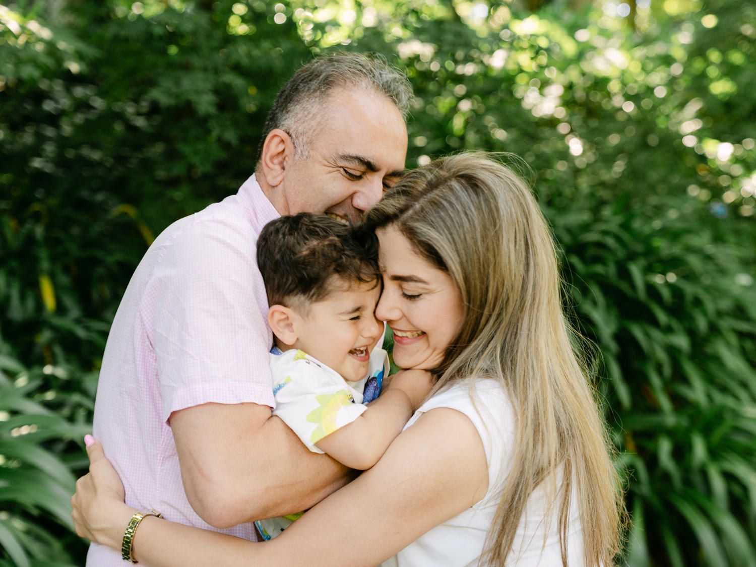 A loving family of three shares a tender embrace outdoors, with lush green foliage in the background.