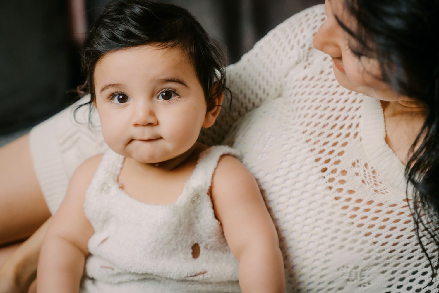 Adorable baby girl in a cream knit romper sitting with her mother, looking curiously at the camera.