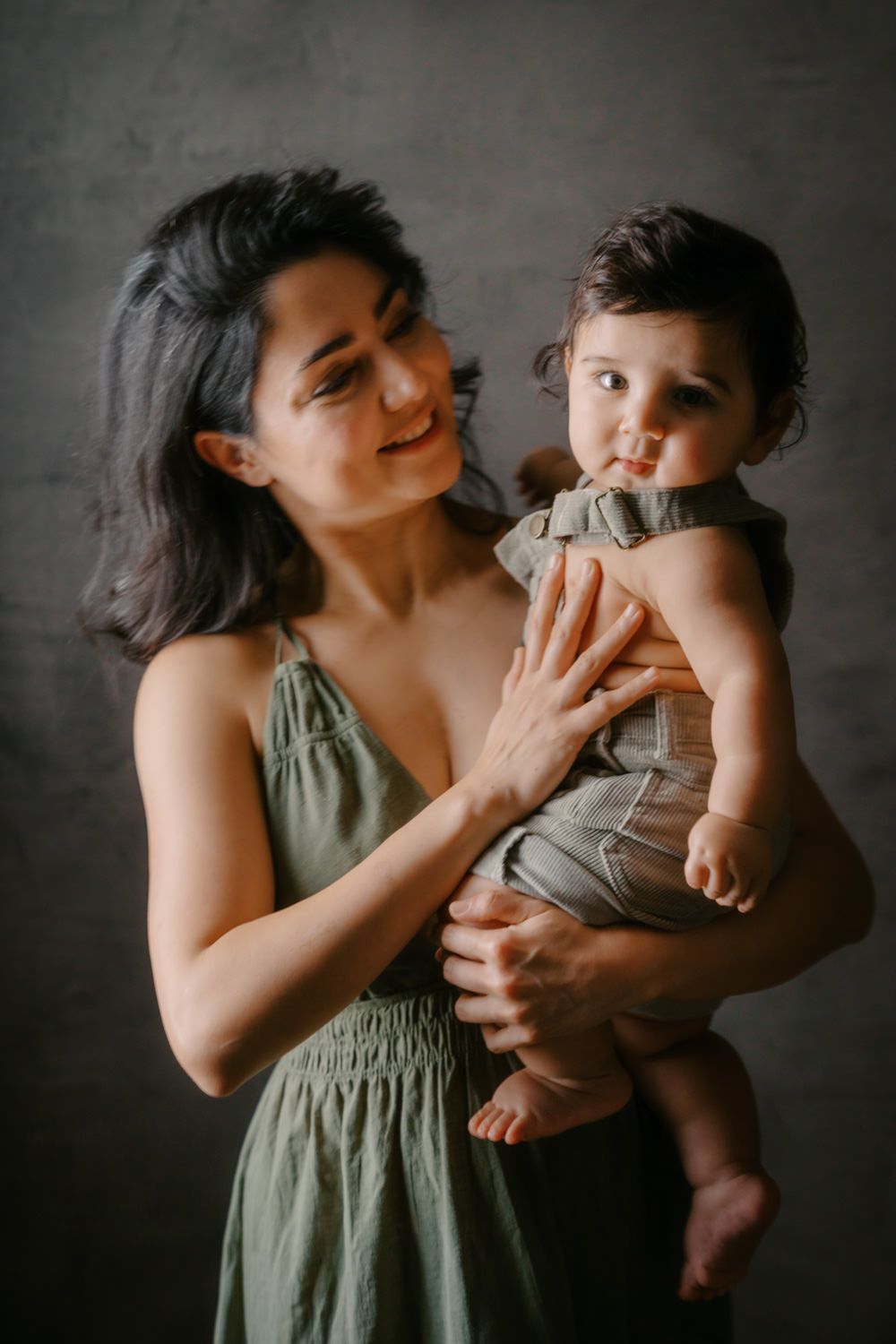 A smiling woman in an olive dress holds a cute baby in a gray romper against a dark studio background.