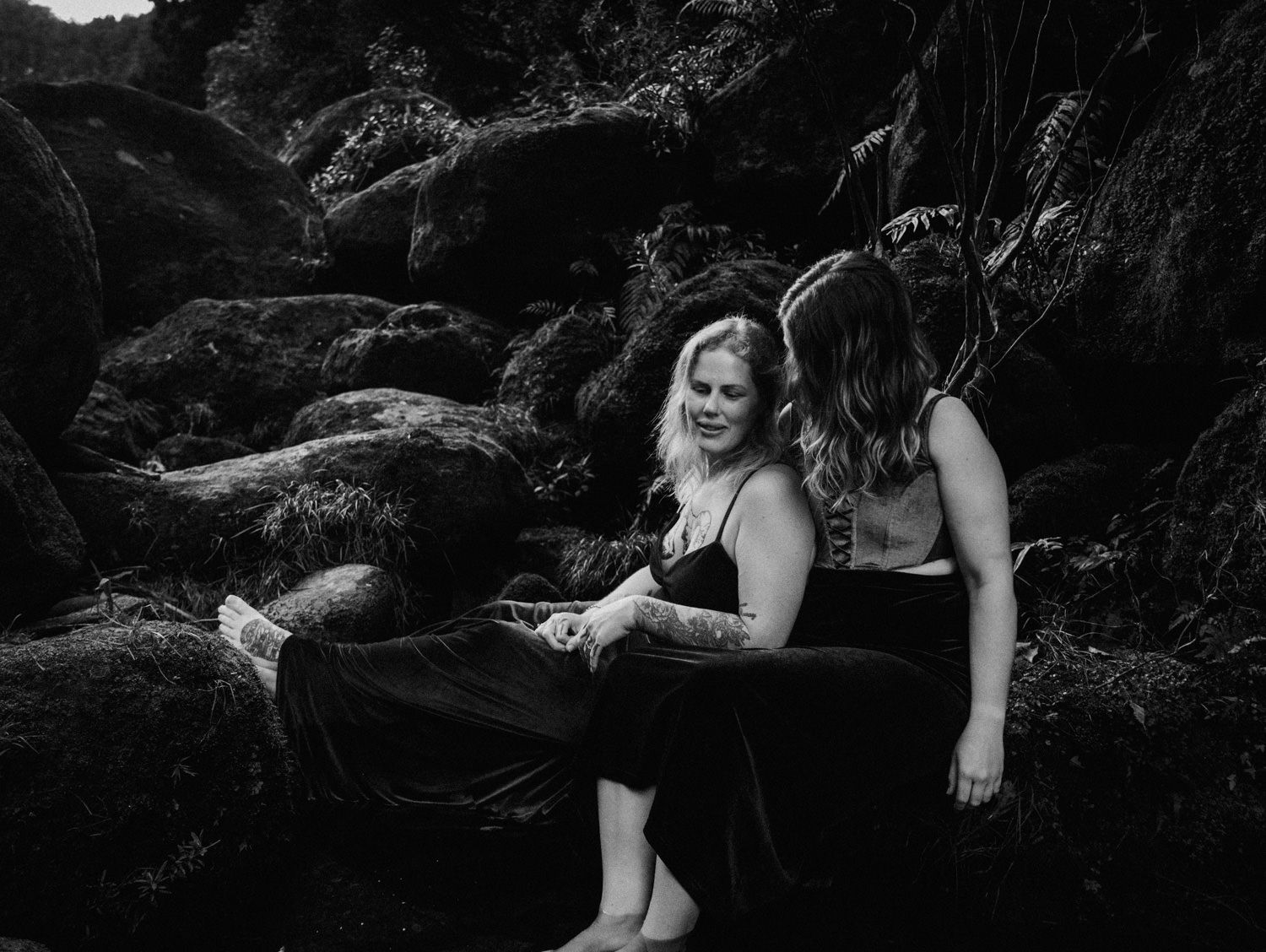 Two women sit together on mossy rocks in a black and white outdoor portrait photo.