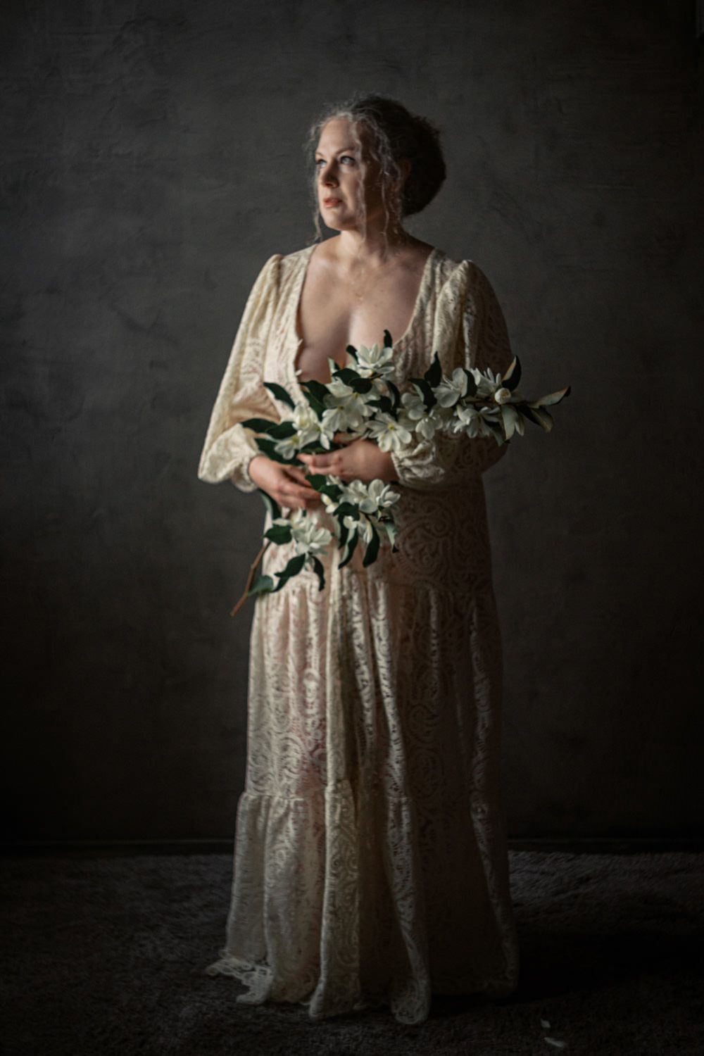 Elegant older woman in lace gown holding white lilies against dark backdrop.