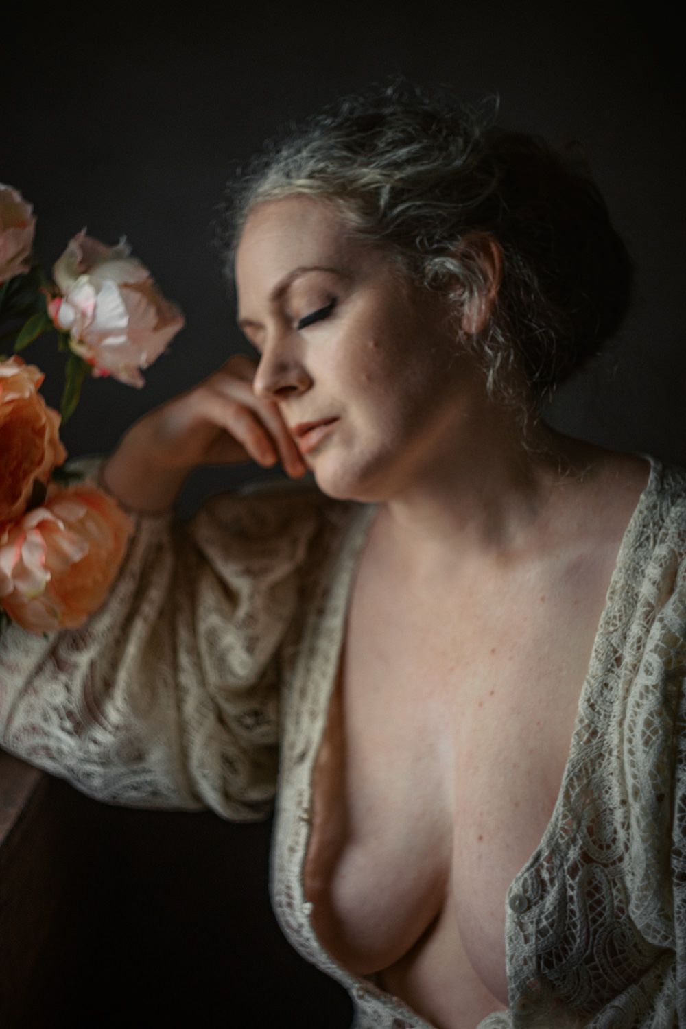 A woman in a lace robe poses with closed eyes beside peach roses in moody studio lighting.