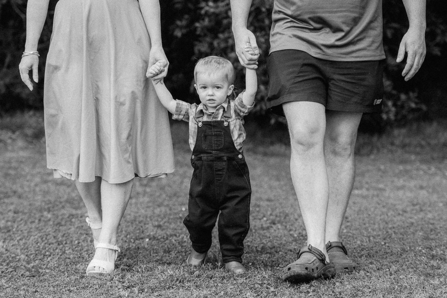 Black and white photo of a toddler in overalls holding hands with parents walking on grass.