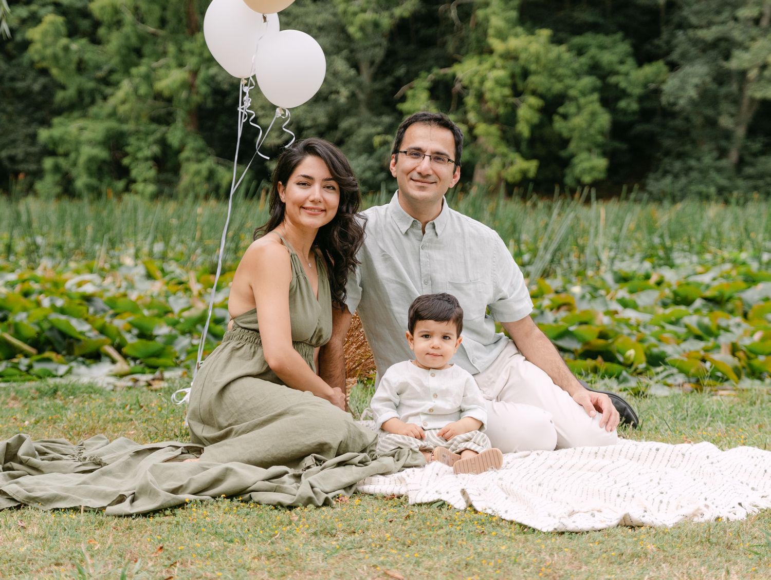 Happy family with parents and baby sitting on blanket in park with white balloons for a milestone photo.