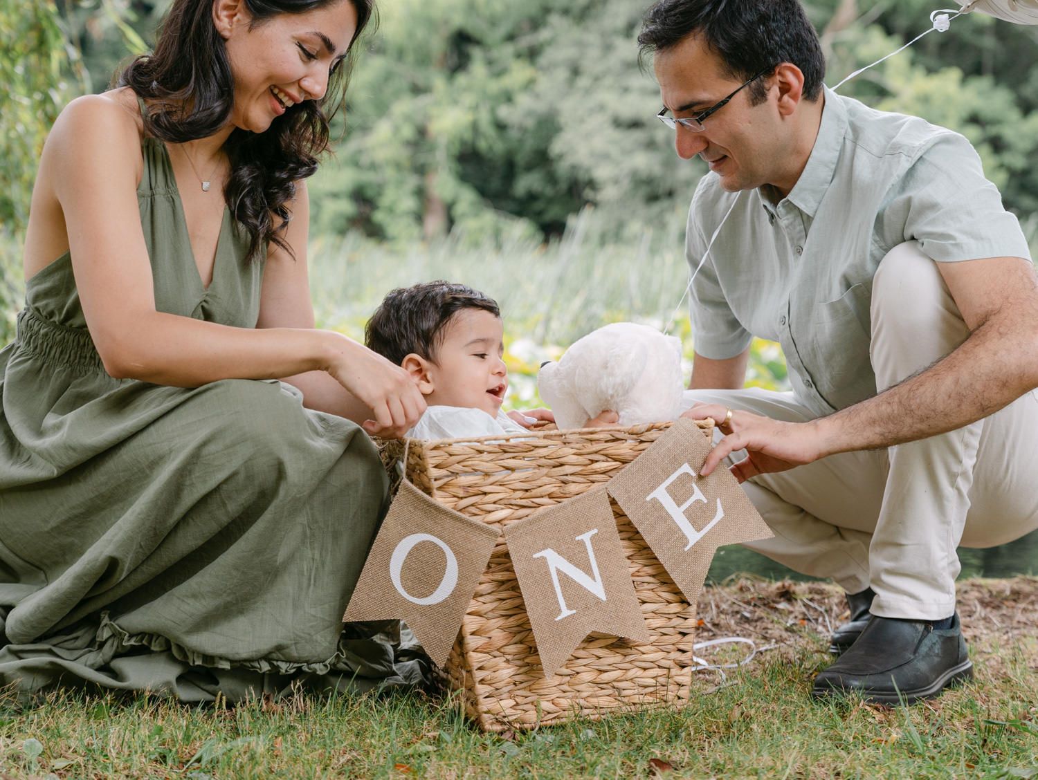Family celebrating baby's first birthday with ONE banner and white bunny in a wicker basket outdoors.