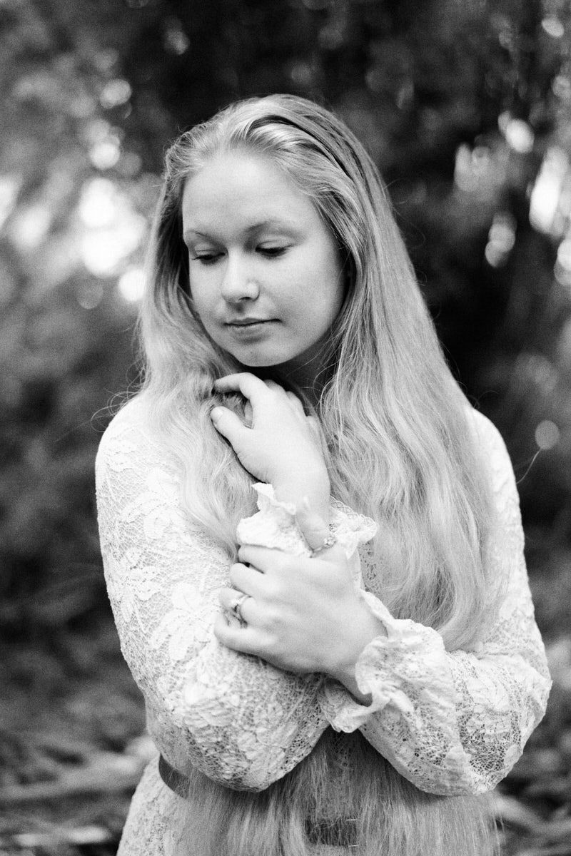 Black and white portrait of a young woman with long blonde hair in a lace dress, eyes closed, arms crossed.