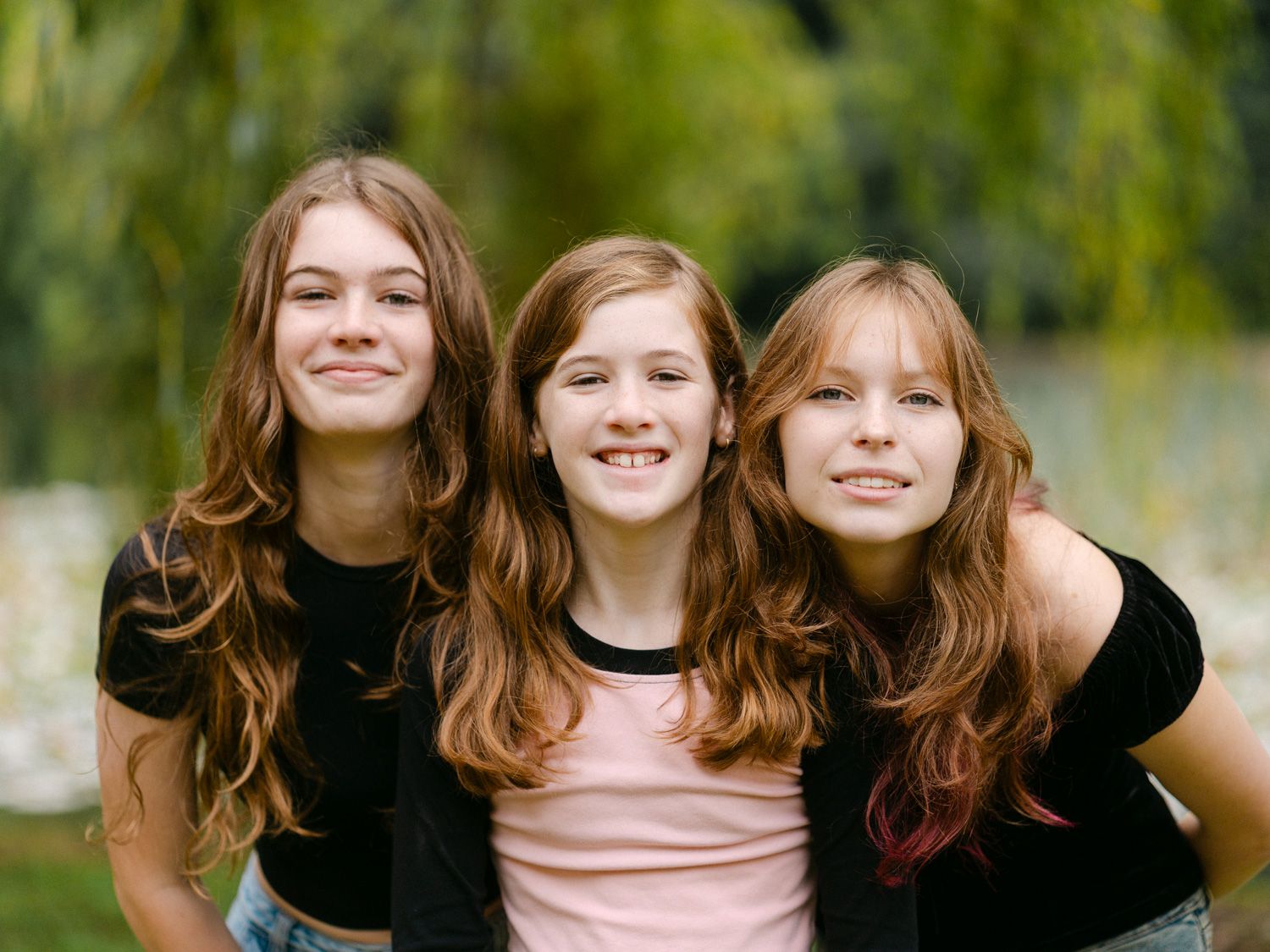 Three smiling teenage girls with long brown hair pose together outdoors with green trees in the background.