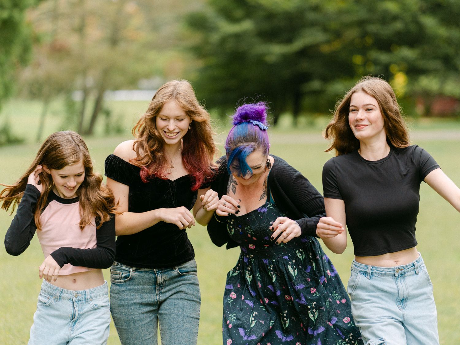 Four young women dancing and laughing together outdoors on green grass, wearing casual tops and jeans.