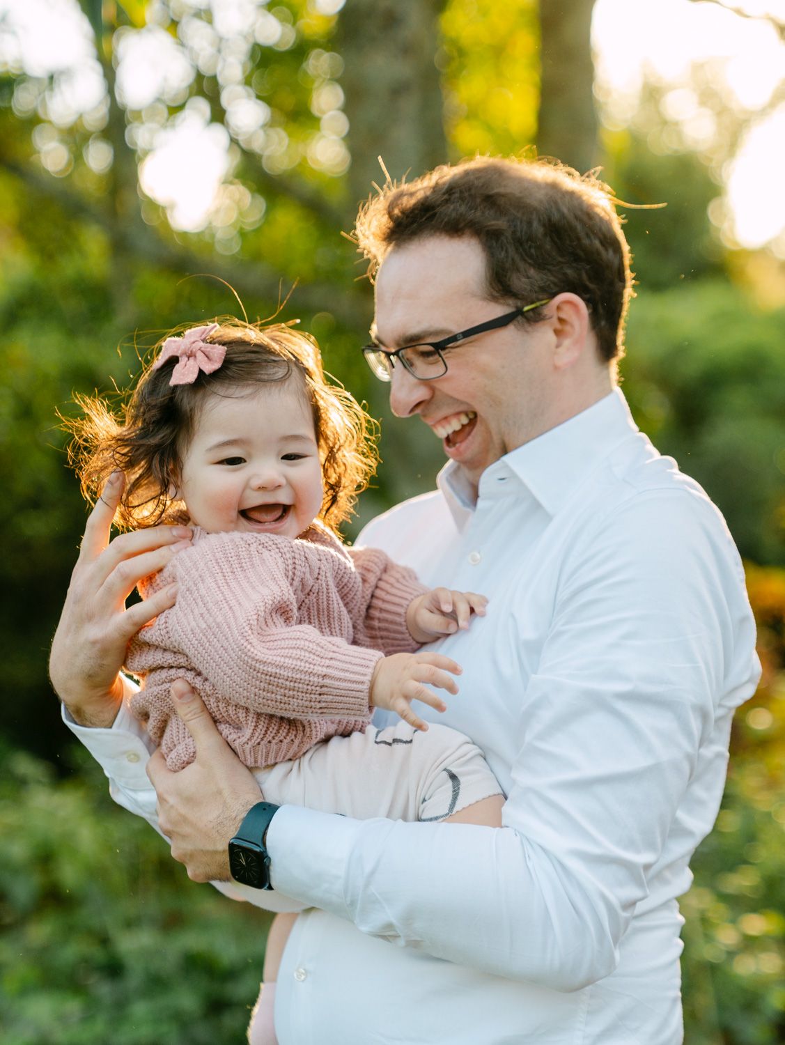 Father laughing while holding smiling baby girl in pink sweater outdoors in golden hour light.