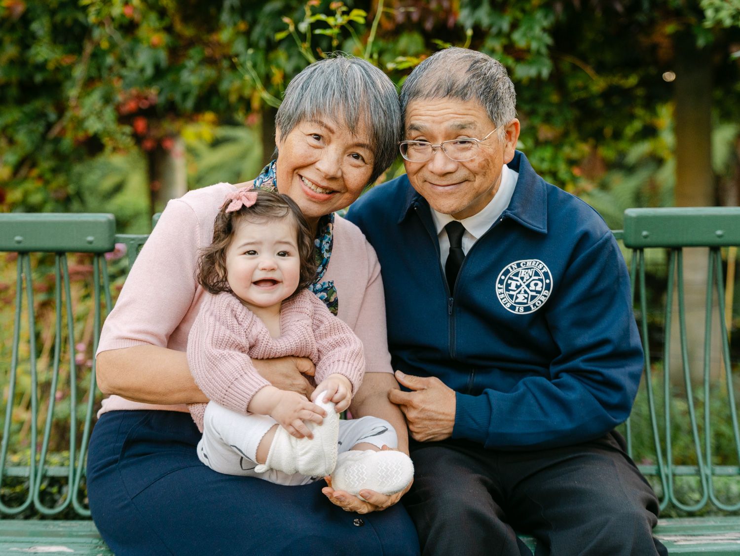 Grandparents sitting on a bench outdoors, smiling with a happy baby girl in a floral outfit.