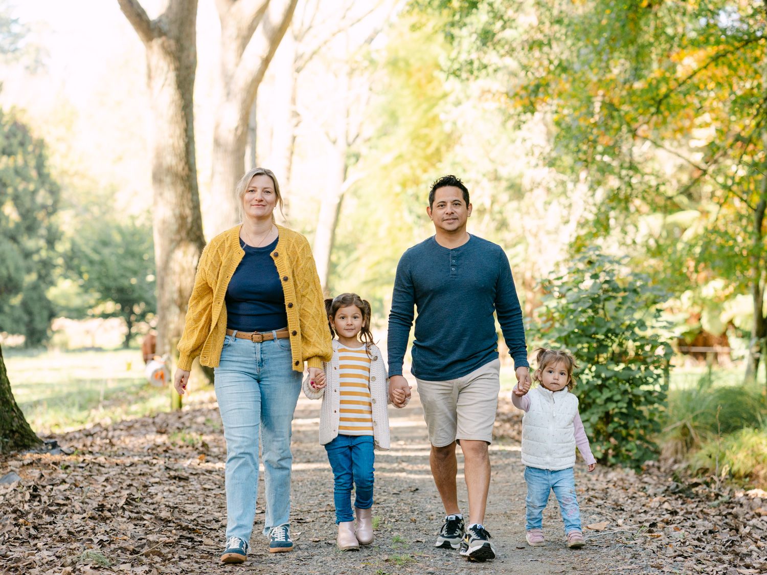 Family of four walking hand-in-hand on a park path surrounded by autumn trees.