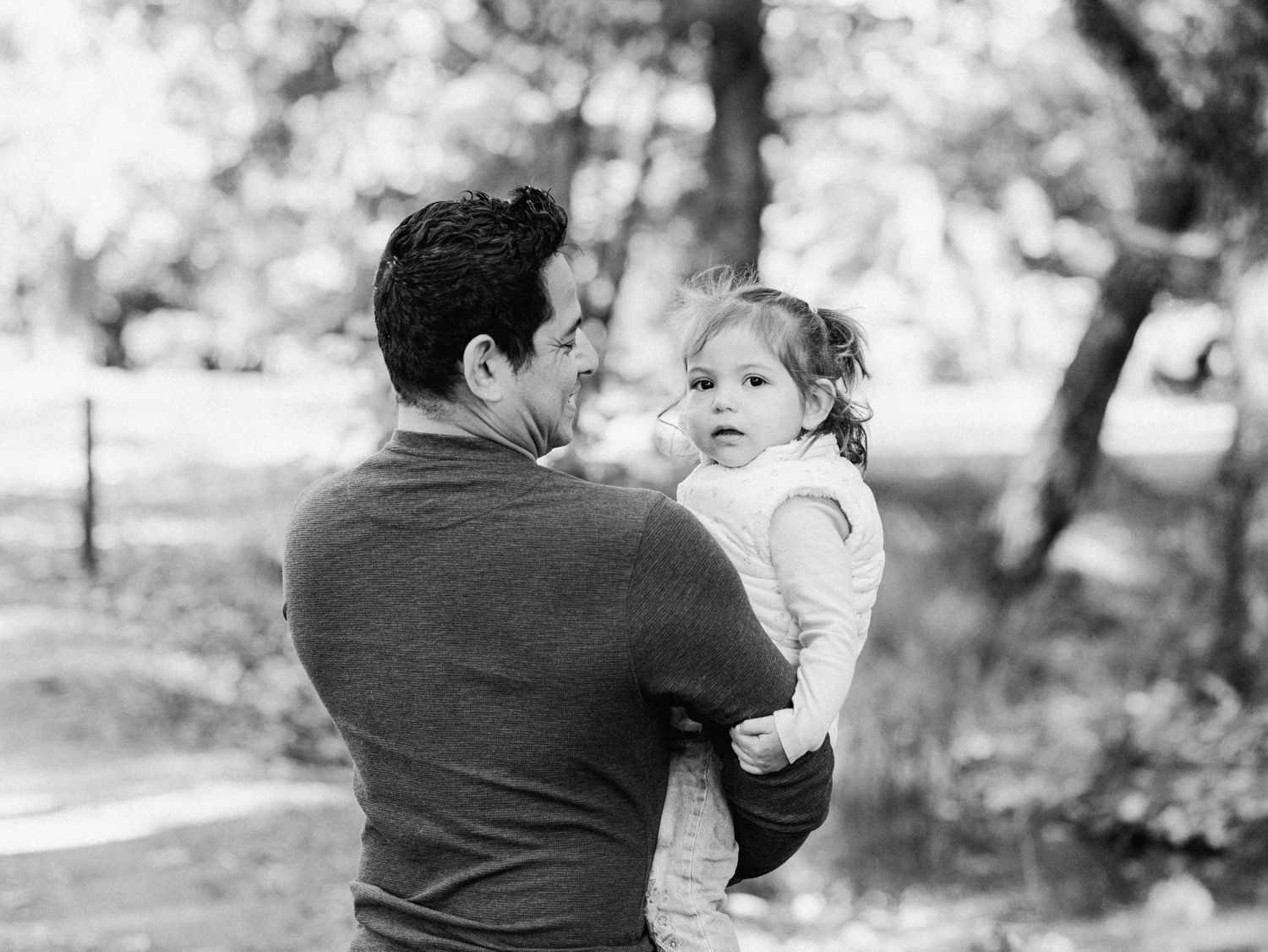 Black and white photo of a father holding his toddler daughter outdoors in a park setting.