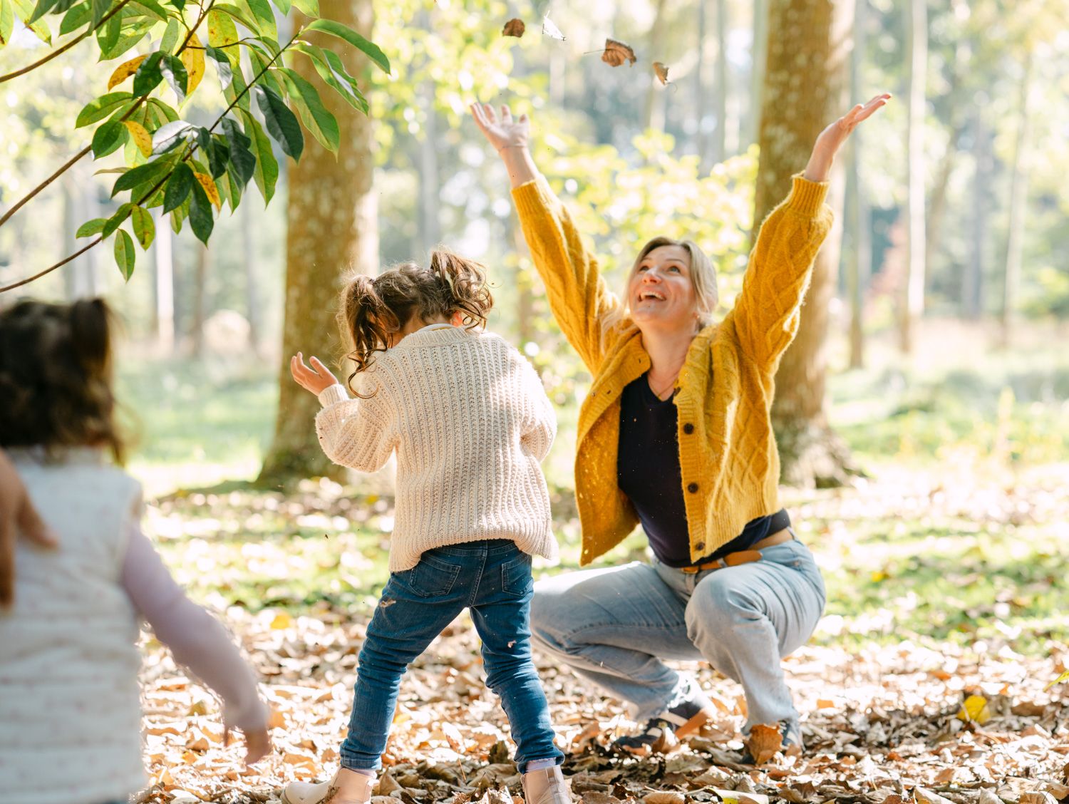 A joyful mother in a yellow cardigan tosses autumn leaves with her children in a sunlit forest.