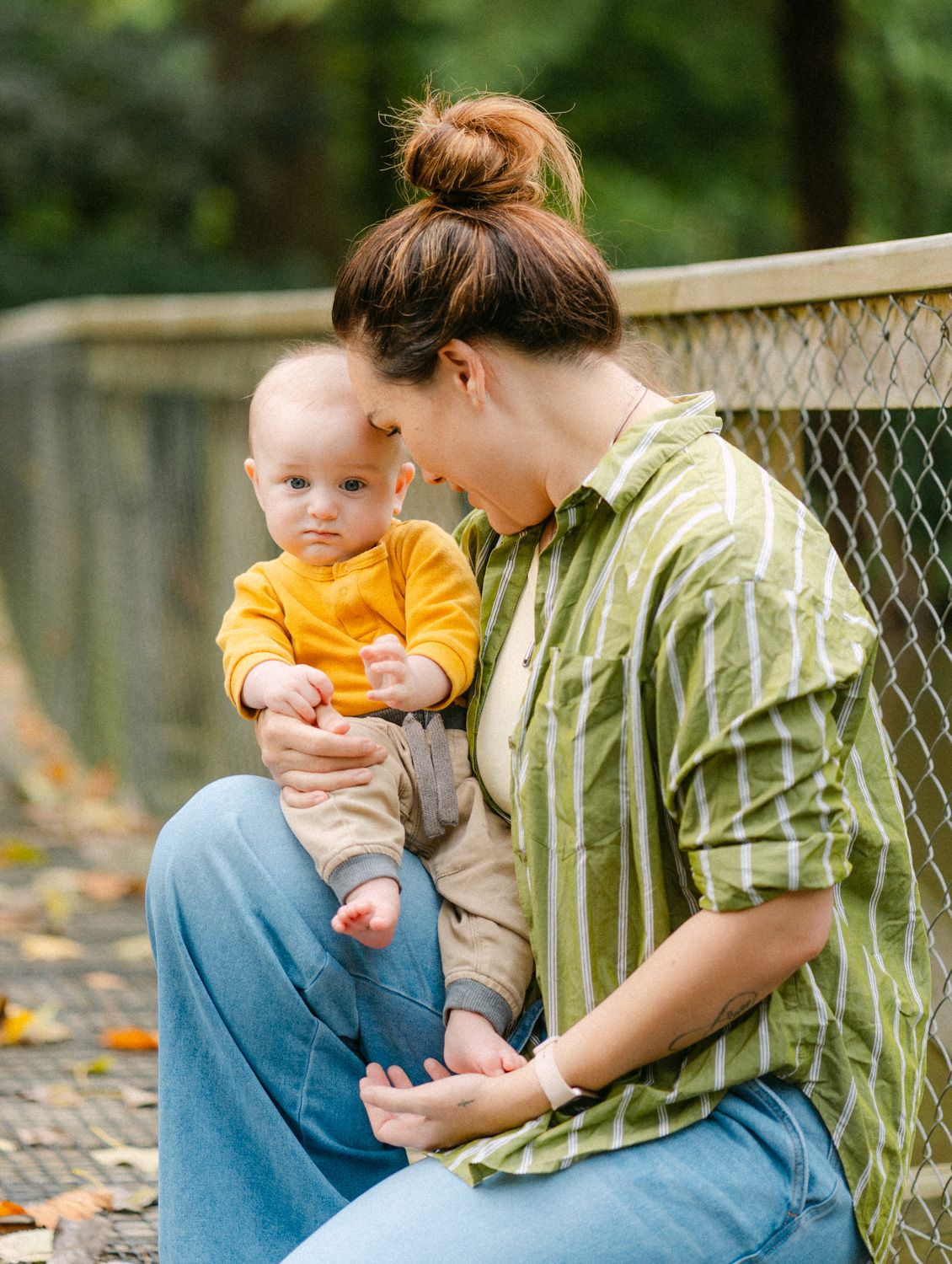 Mother in green striped shirt holds baby in yellow outfit on outdoor bridge surrounded by autumn leaves.