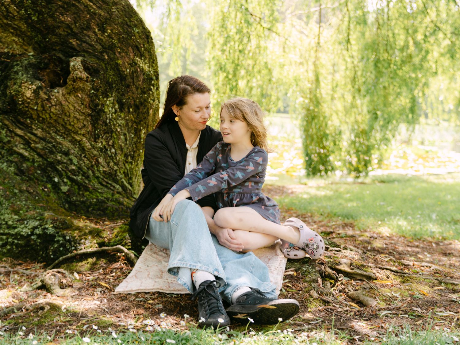 A mother and daughter sit together against a mossy tree in a sunlit park, sharing a warm embrace.