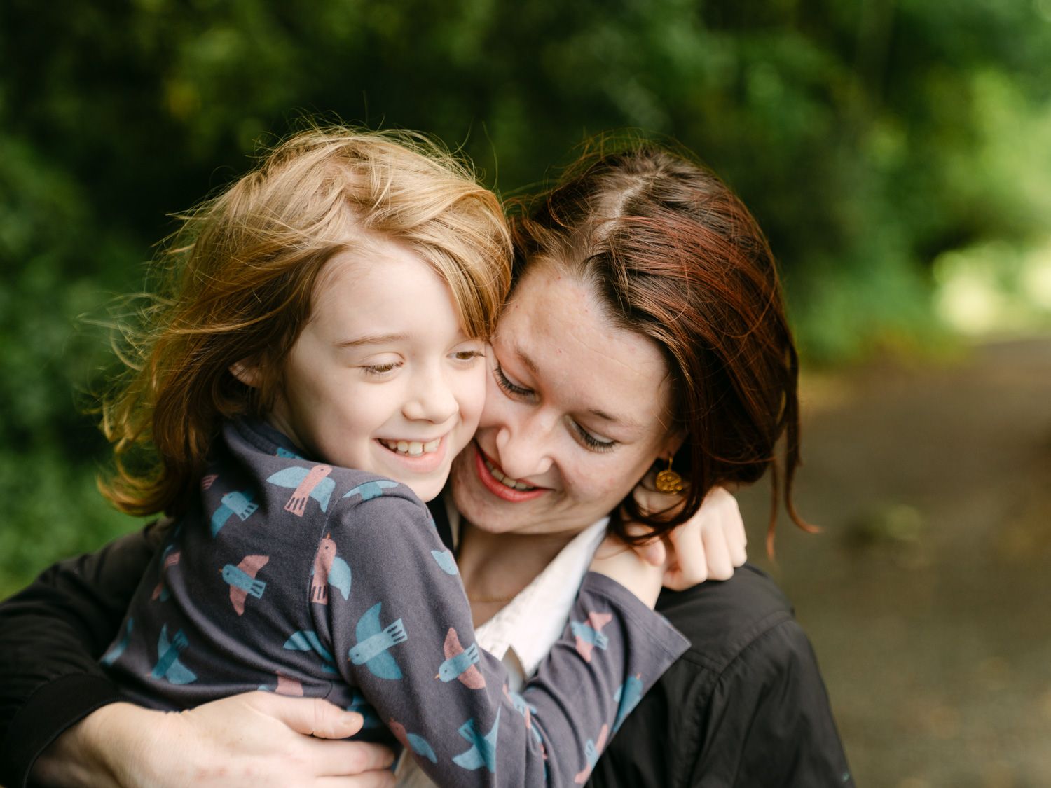 A joyful redhead child hugs a smiling brunette woman outdoors, sharing a warm embrace in a lush green setting.