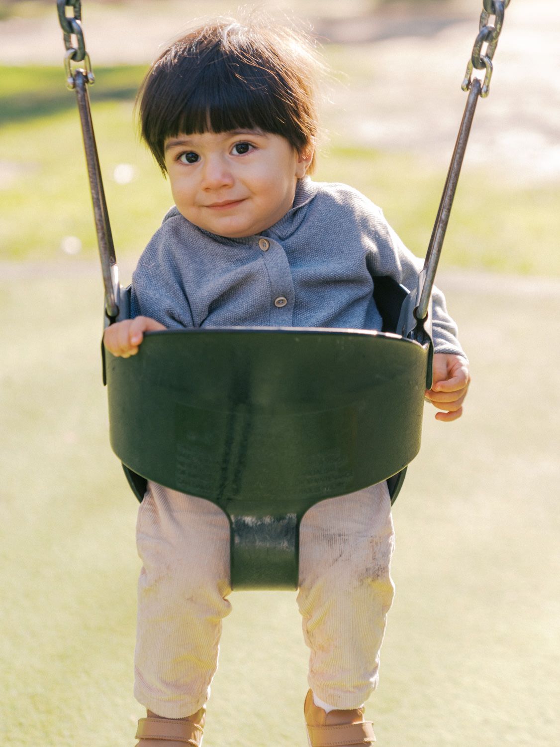 Adorable toddler sitting in a green bucket swing at a playground, smiling at the camera.