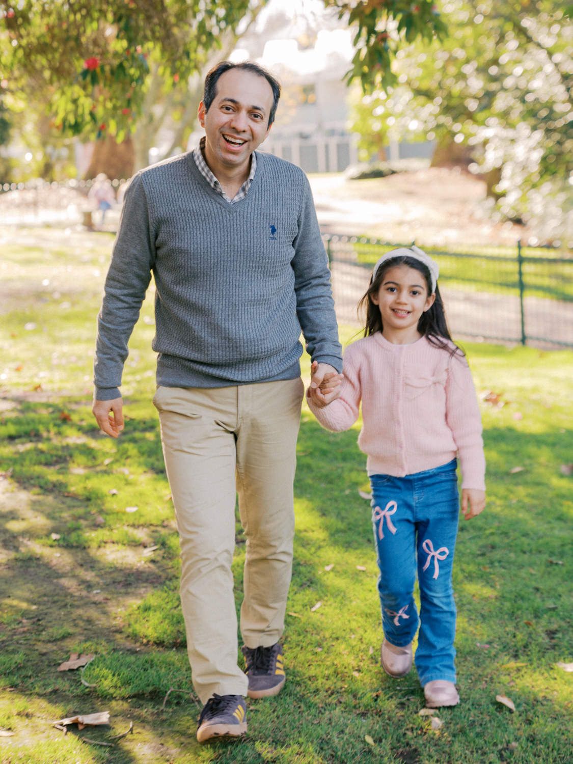 A man and young girl walk hand-in-hand through a sunny park, both smiling, wearing casual clothes.