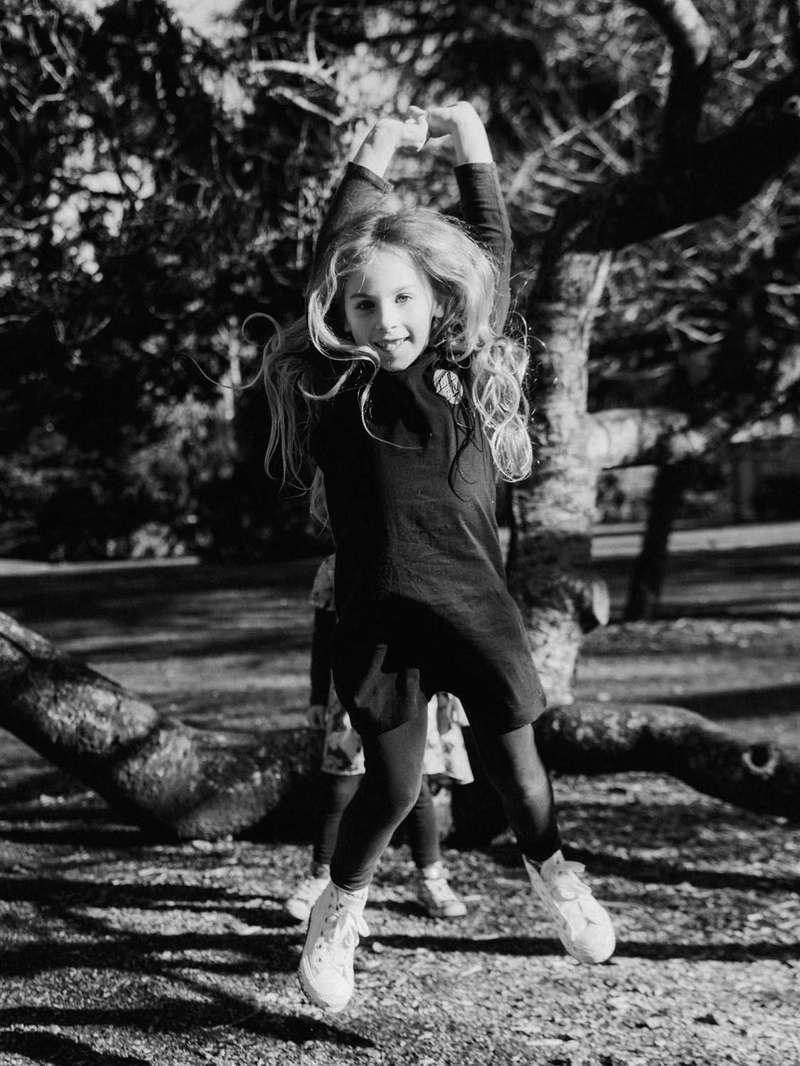 A joyful girl jumps in the air with arms raised in a black-and-white photo taken outdoors near trees.