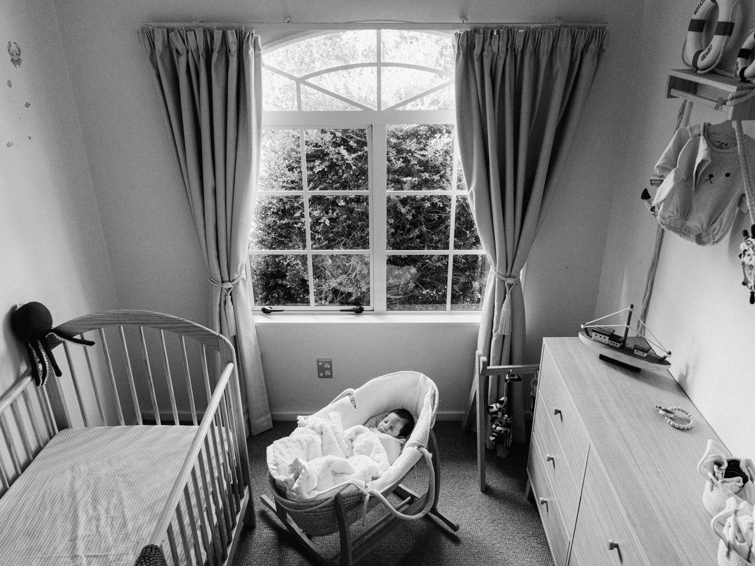 Black and white photo of a cozy nursery room with a crib, Moses basket, and dresser by a window.