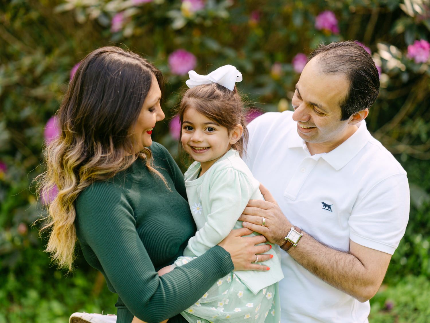A happy family of three poses outdoors, with parents holding their young daughter in a lush garden setting.
