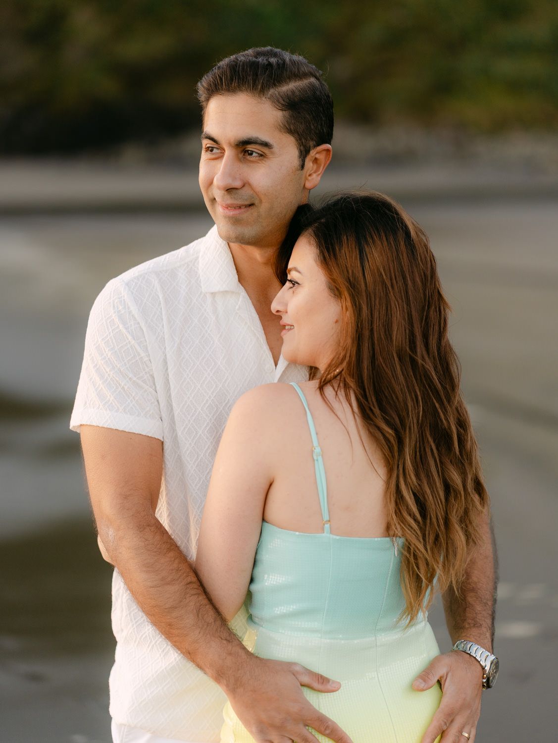 Couple embracing on a beach at sunset, man in white shirt holding woman in mint green dress.