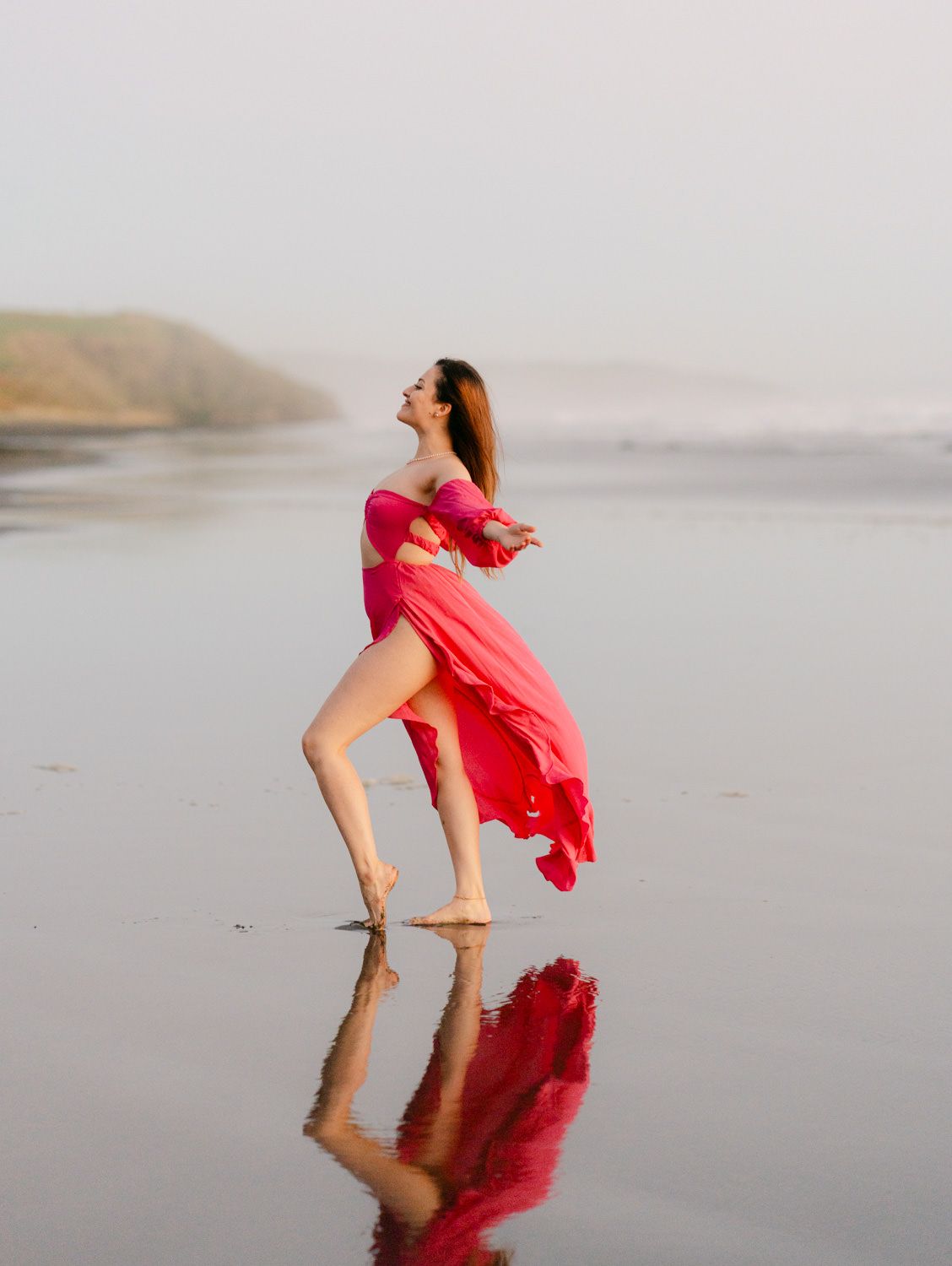Woman in flowing red dress dancing barefoot on reflective wet beach with misty coastal cliffs in background.