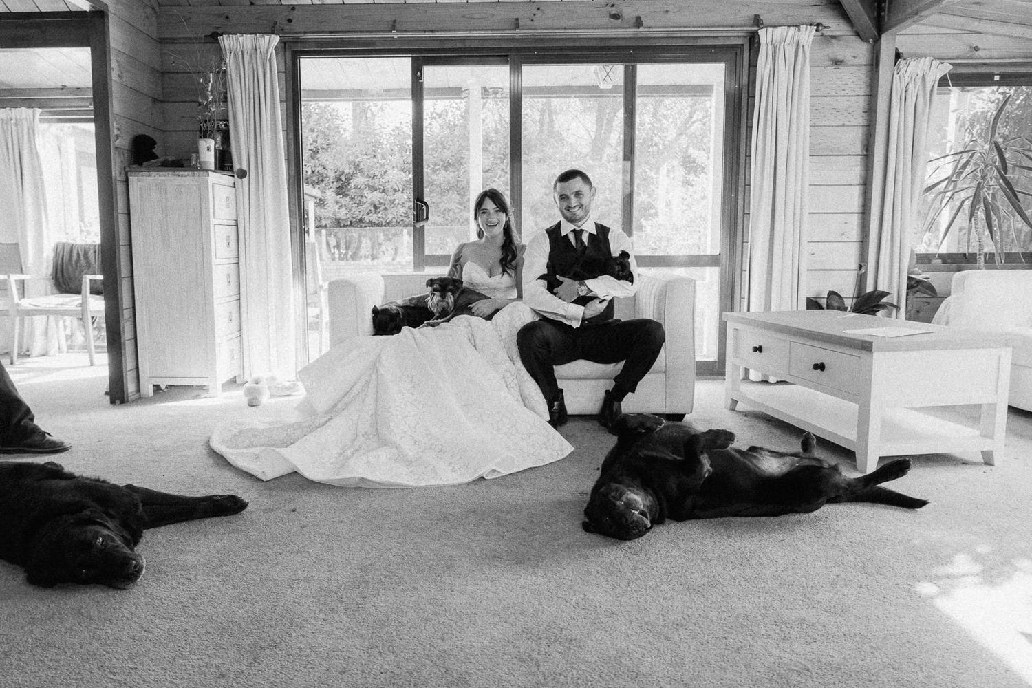 Black and white wedding photo of bride and groom sitting together indoors with two black Labrador dogs lying on the floor.