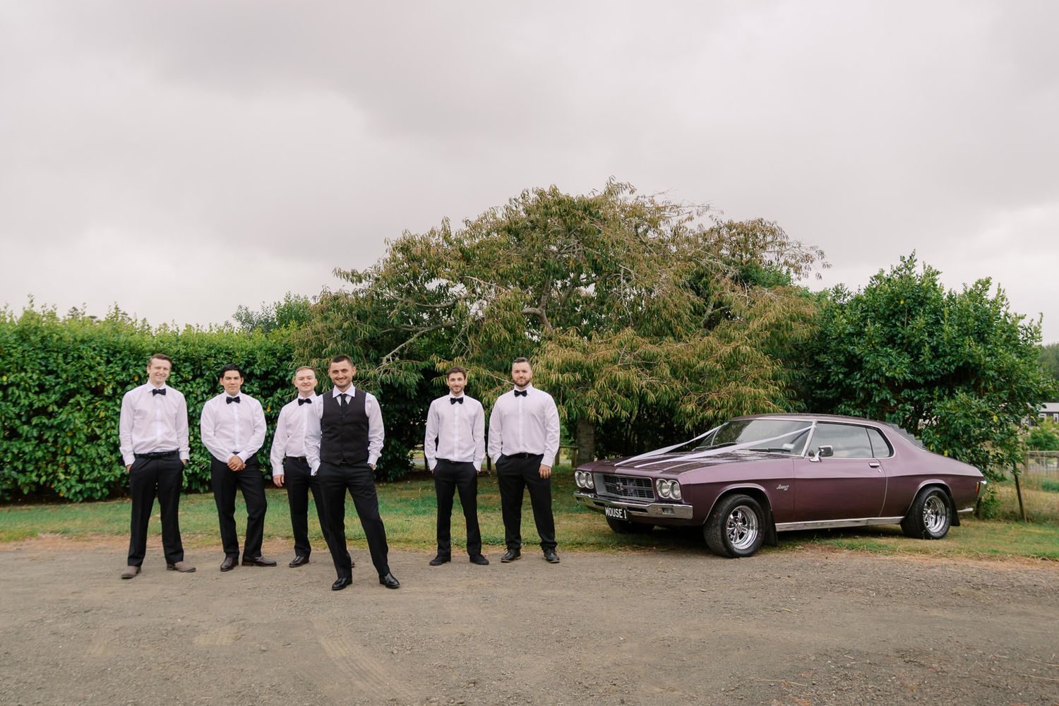 Groom and groomsmen in black pants and white shirts with bow ties standing beside a classic maroon car at a wedding.