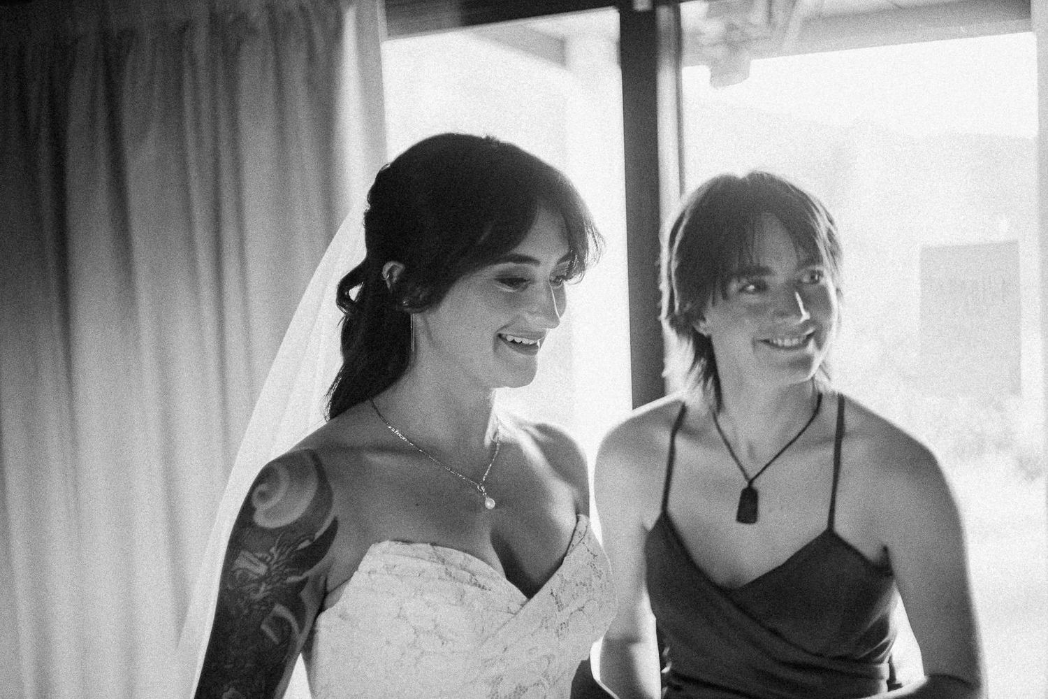 Black and white photo of a tattooed bride in a lace gown and veil smiling with a bridesmaid.