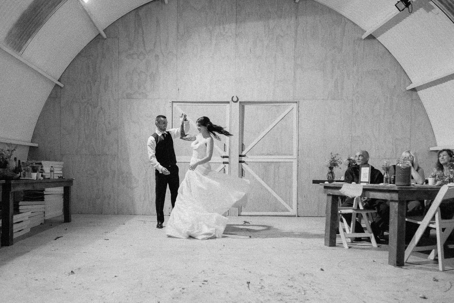 Black and white photo of a bride and groom sharing their first dance in a rustic barn venue with wooden walls.