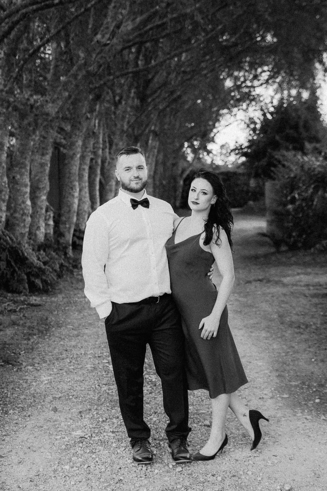 Elegant couple posing on a tree-lined path in black and white, man in bow tie and woman in slip dress.
