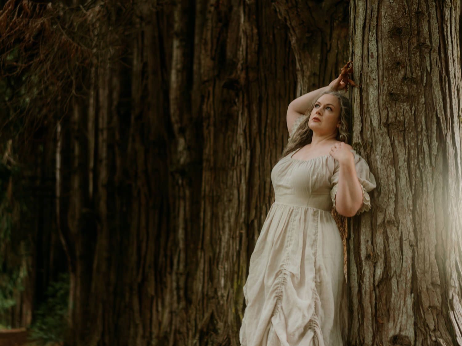 Woman in white dress leaning against large tree trunk in forest, looking upward dreamily.