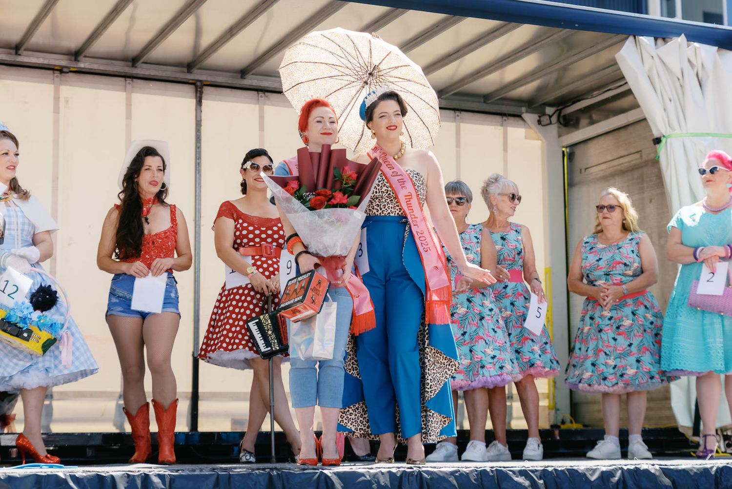 Women in vintage 1950s-style dresses on stage at a retro pin-up contest, one holding umbrella and bouquet.