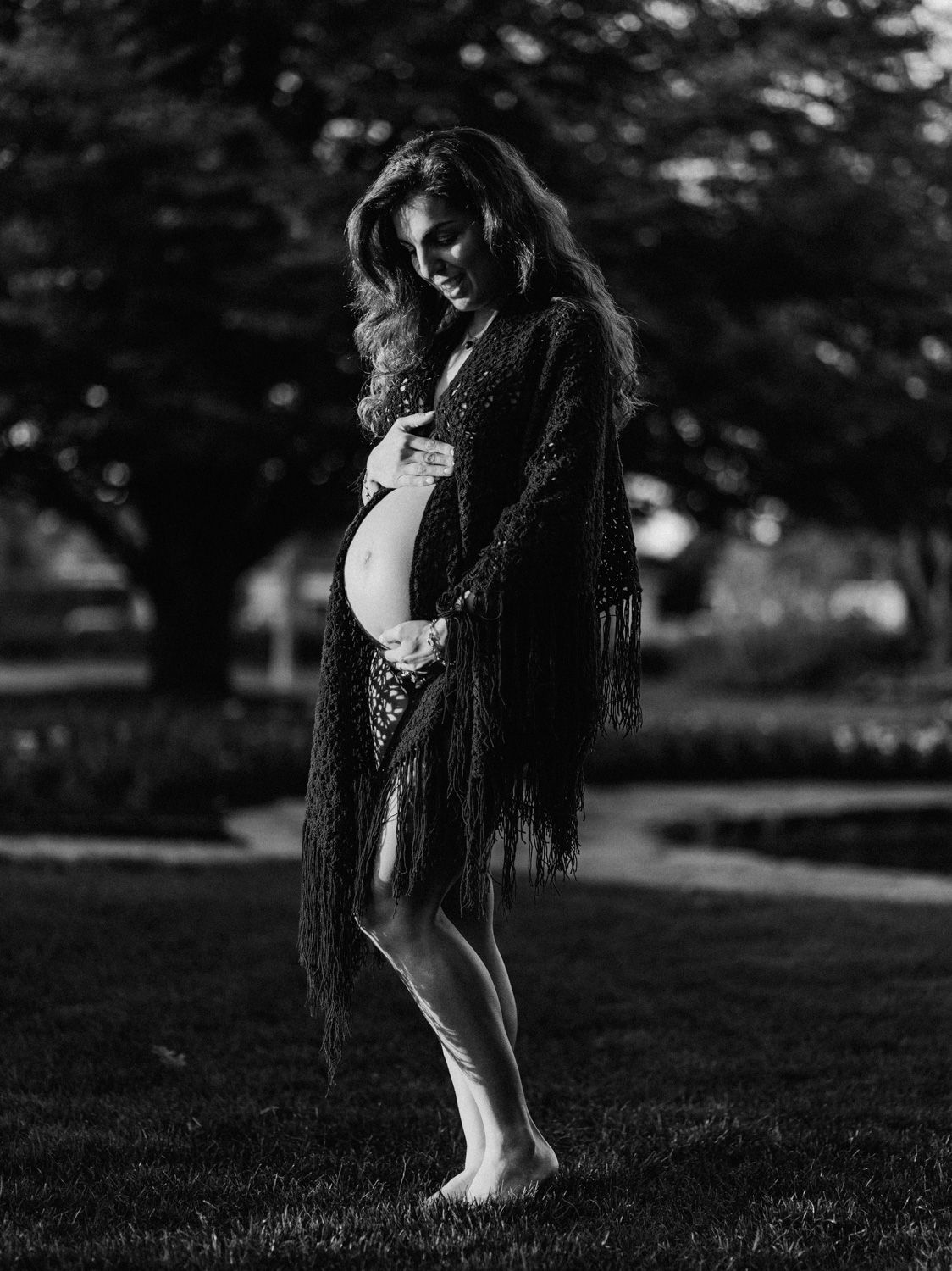 Black and white maternity photo of a woman in a fringed kimono cradling her baby bump outdoors.
