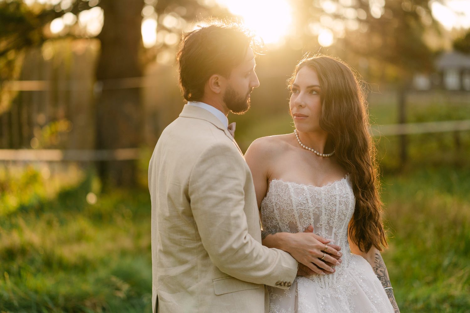 Couple in wedding attire embracing at golden hour sunset in lush green garden setting.