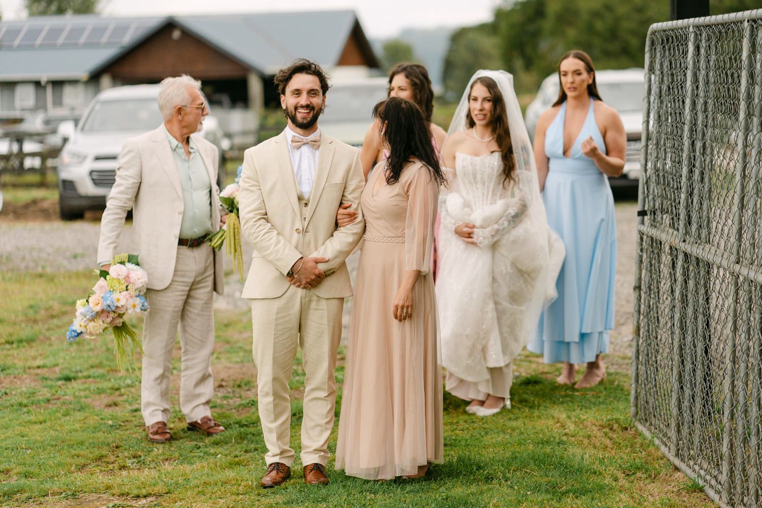 Wedding party walking outdoors on grass, groom in tan suit with bride in white gown, guests in blush and blue dresses.