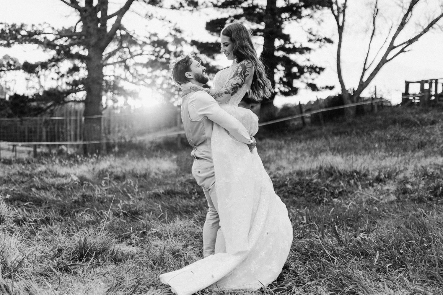 Black and white photo of groom lifting bride in a romantic embrace outdoors at sunset.