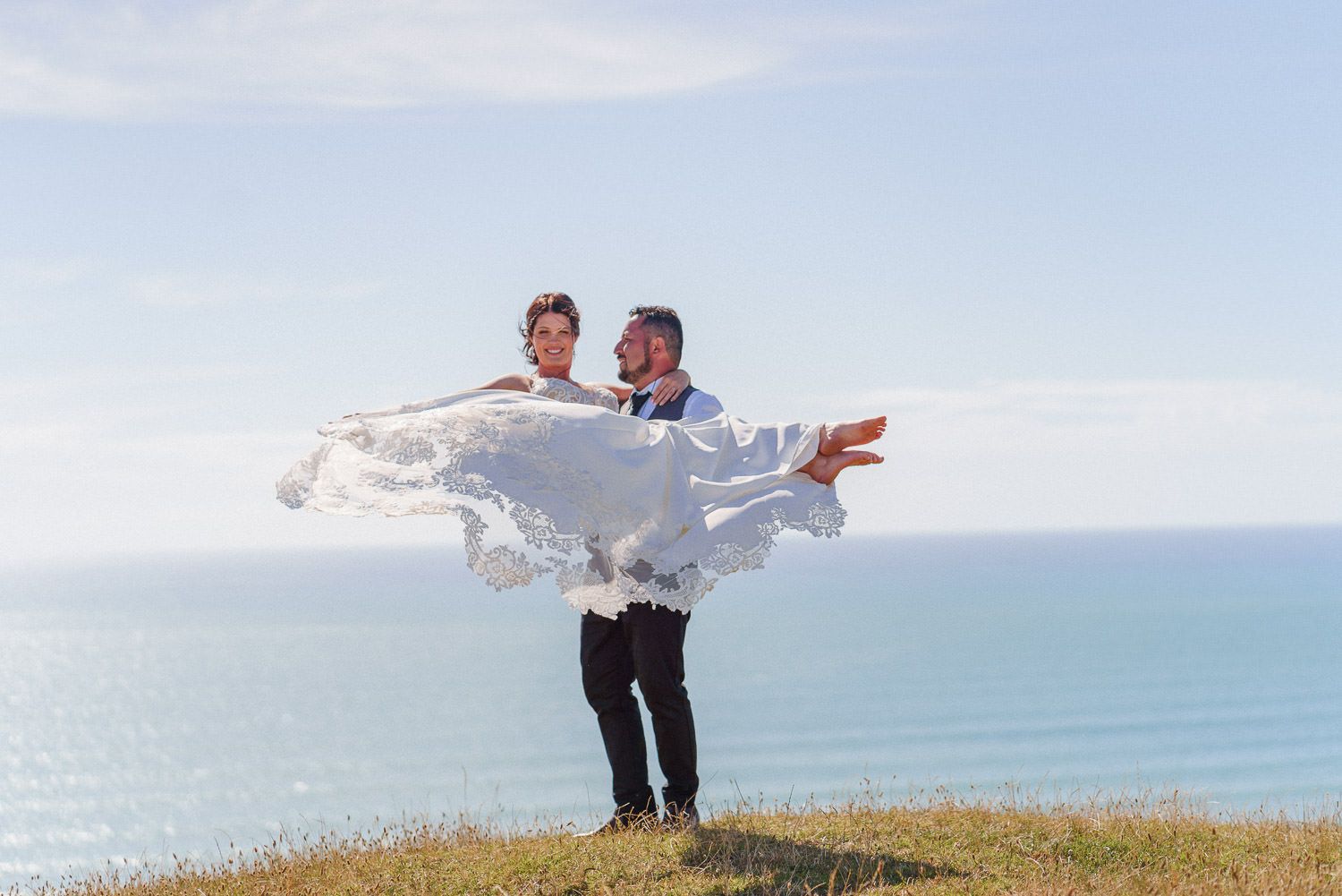 Groom carries bride in flowing white dress on coastal cliff with ocean backdrop on their wedding day.