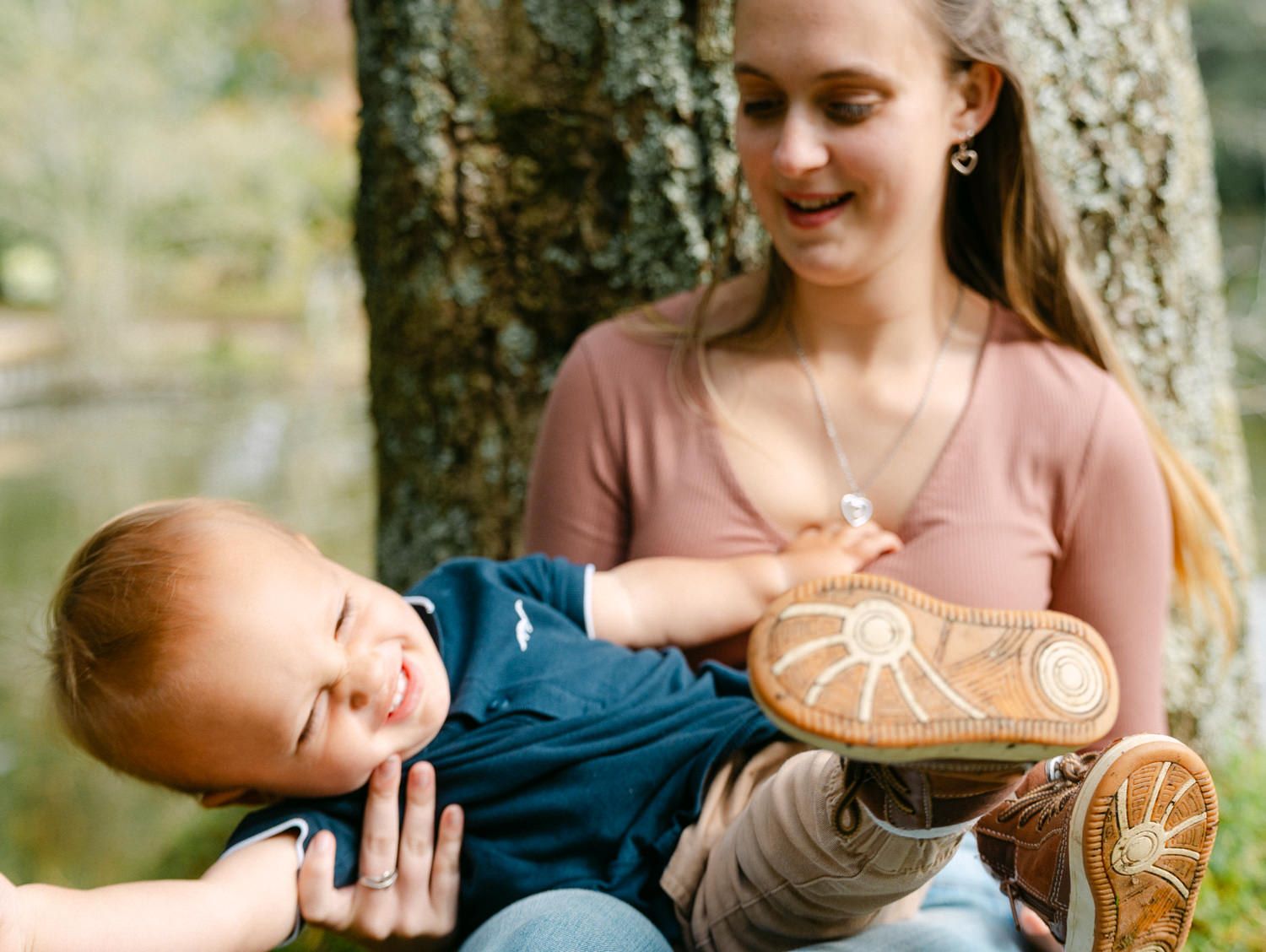 A young woman smiles while playfully holding a laughing baby outdoors near a tree.
