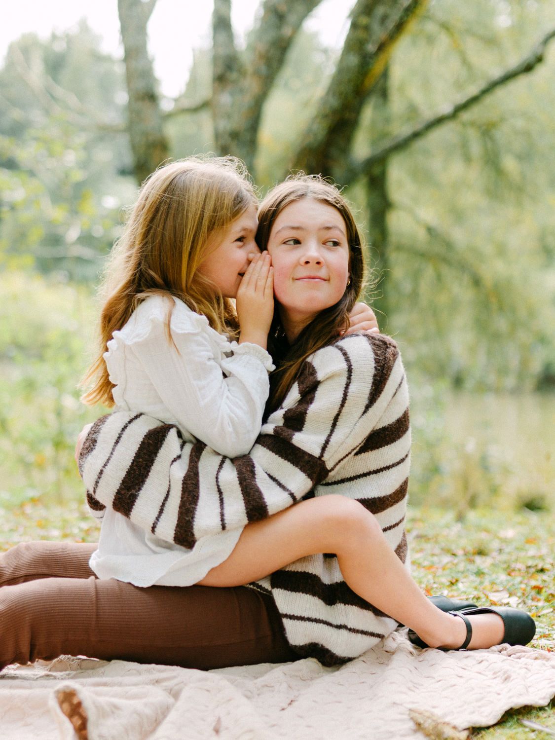 Two young girls sit outdoors on a blanket, one whispering in the other's ear, wearing striped sweater.