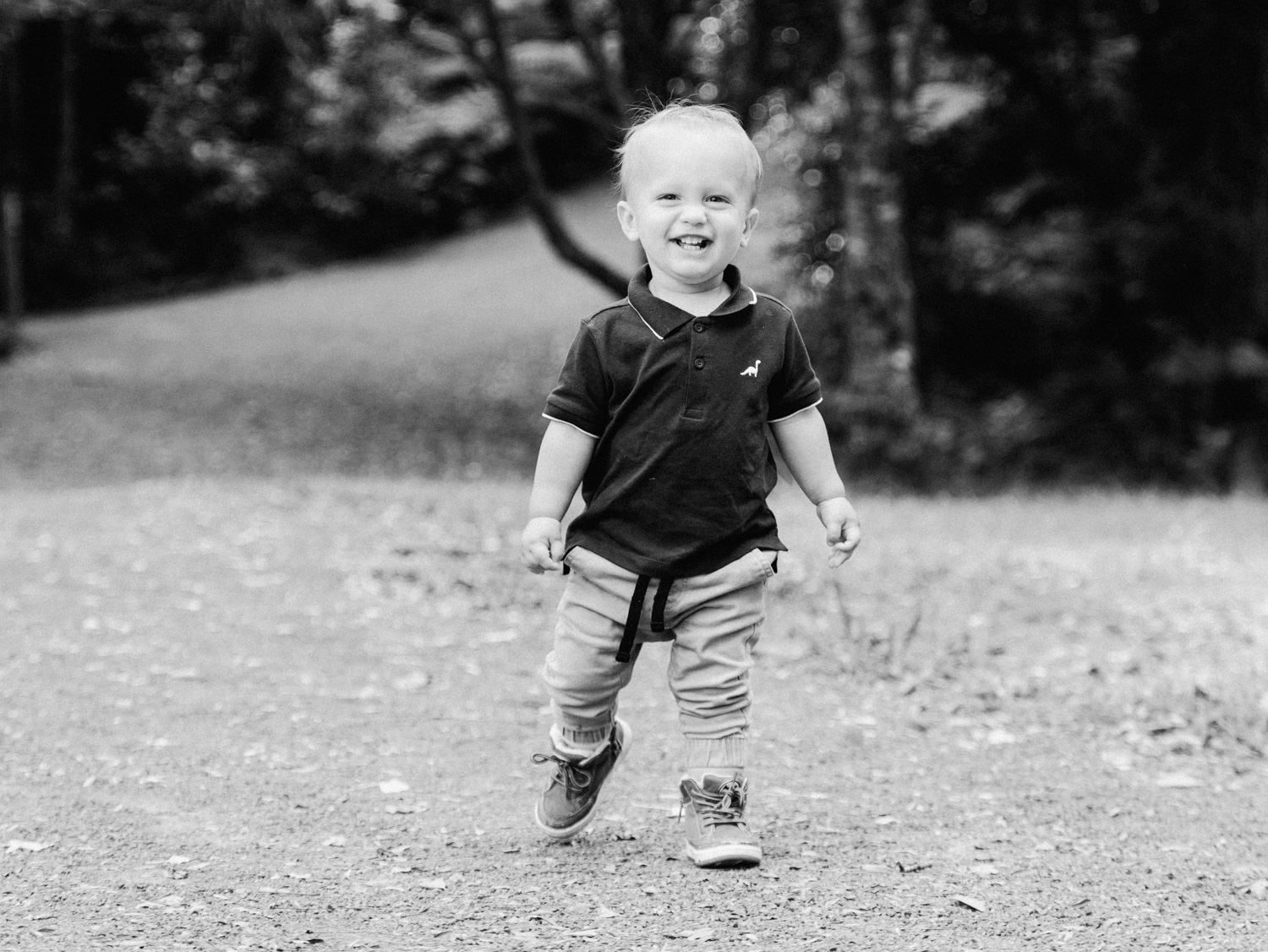 Black and white photo of a happy toddler boy walking on a path outdoors, smiling brightly.